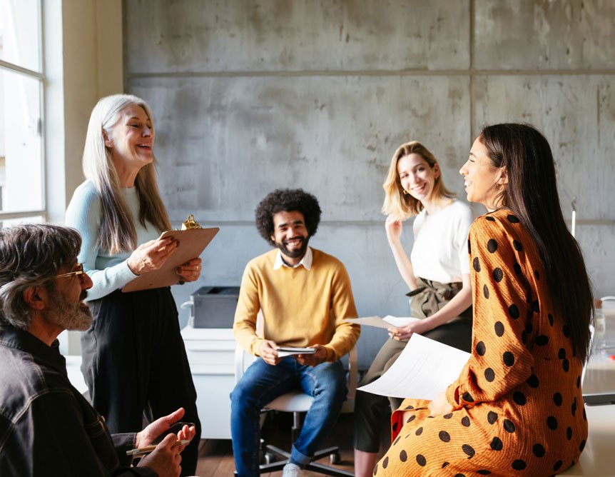 Five people sit and stand in an office, smiling and talking together in a relaxed meeting.