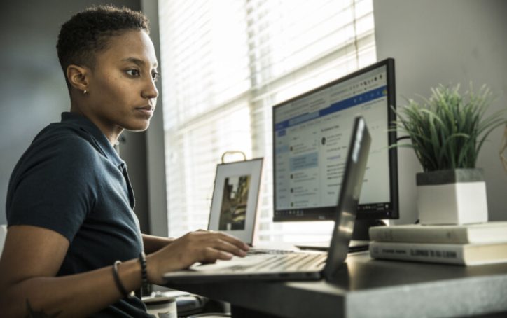 Person working at a desk with a laptop and monitor, next to a window with blinds and a potted plant.