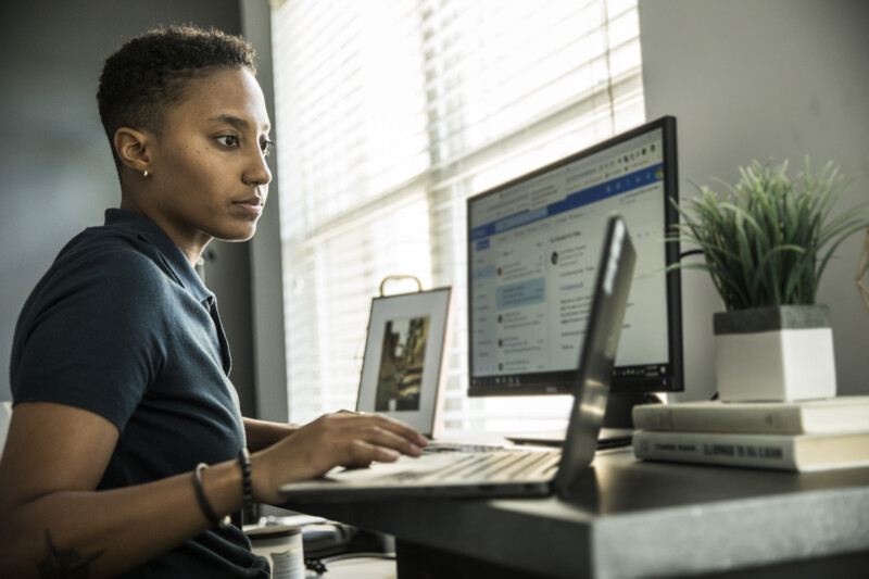 Person working at a desk with a laptop and monitor, next to a window with blinds and a potted plant.