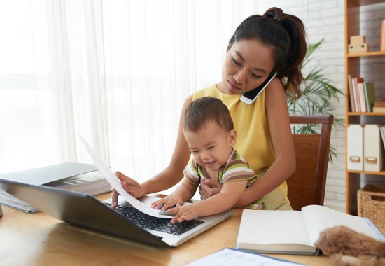 Woman working on a laptop while holding a baby, talking on the phone, and looking at documents.