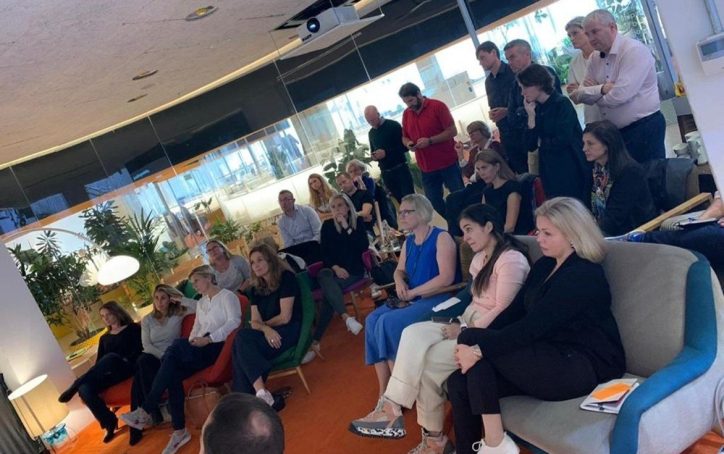 A group of people sit and stand in a modern office, attentively listening to a presentation.