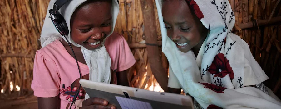 Two girls wearing headscarves smile while looking at a tablet; one wears headphones.