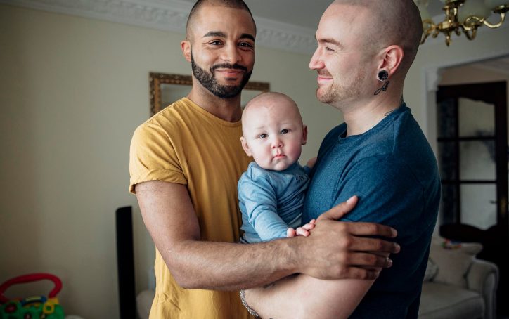 Two men smiling and holding a baby together in a cozy living room.