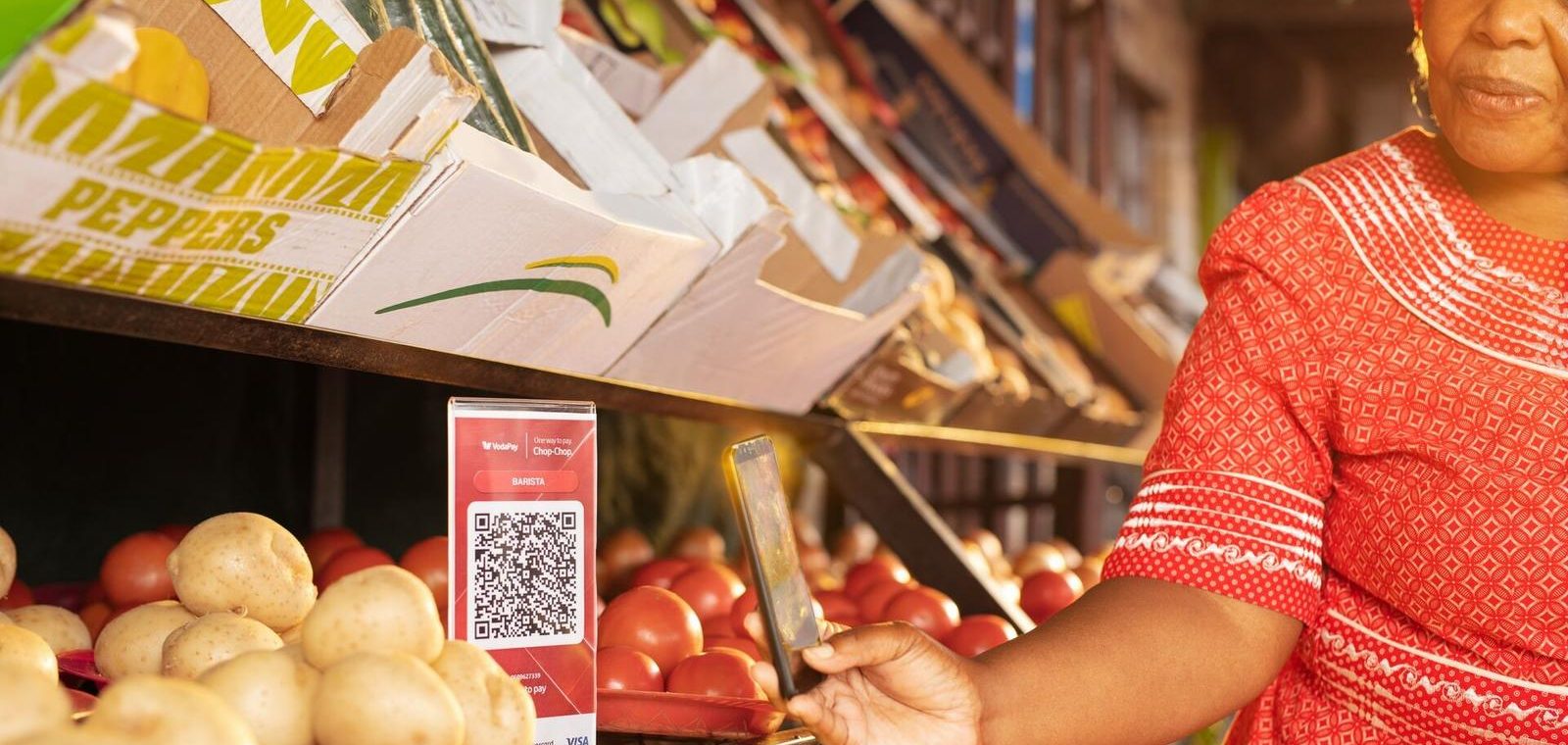 Woman in red dress scans a QR code with her phone at a market stall selling potatoes and tomatoes.