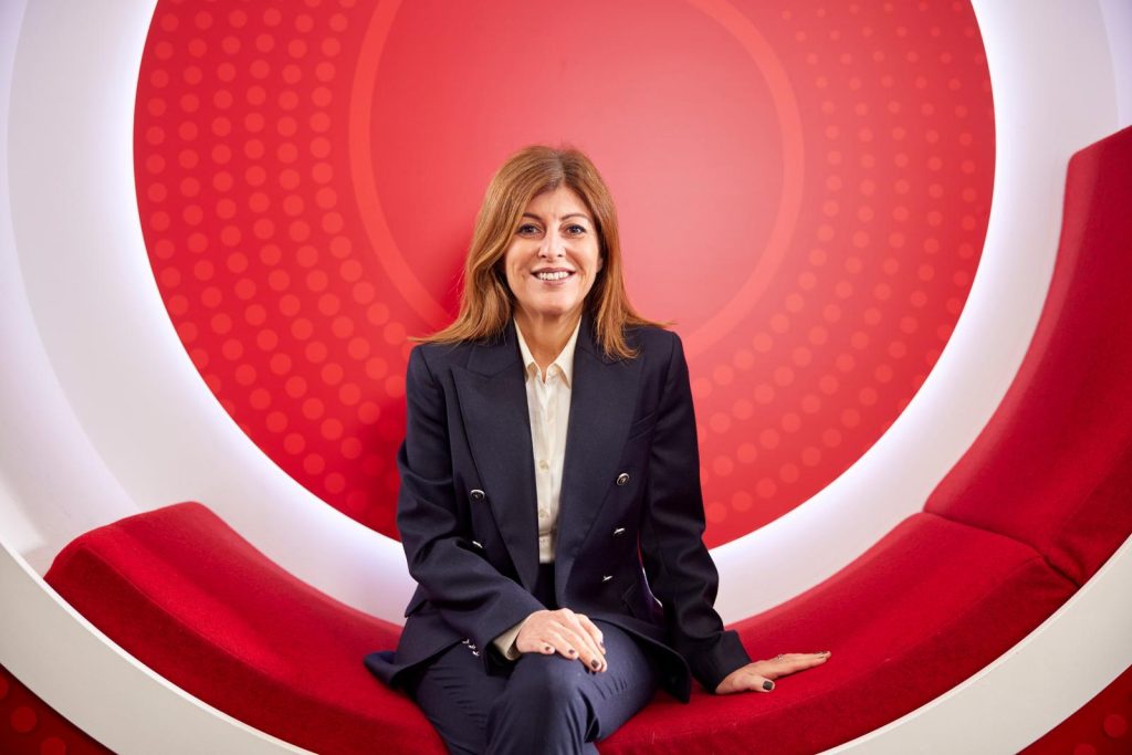 A woman in a dark suit sits on a curved red bench with a circular red and white background.