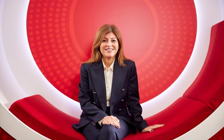 A woman in a dark suit sits on a curved red bench with a circular red and white background.