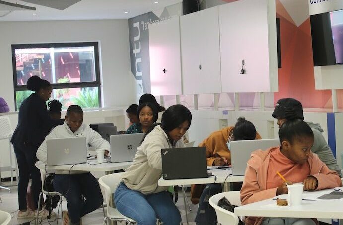 Students sitting at tables working on laptops and writing in notebooks in a modern classroom setting.