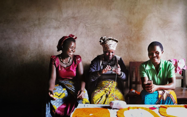 Three women in colorful clothing sit on a bench indoors, smiling and using mobile phones, with crafts on the table.
