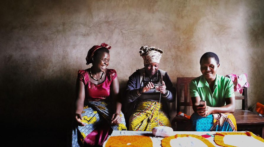 Three women in colorful clothing sit on a bench indoors, smiling and using mobile phones, with crafts on the table.
