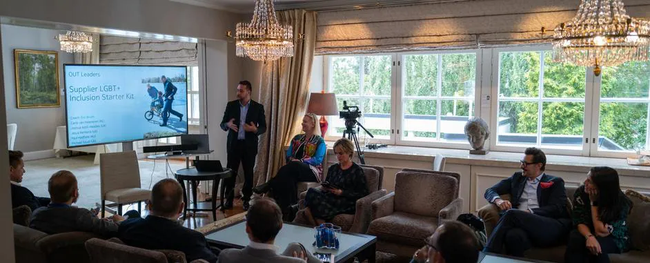 People attend a business meeting in a bright room; a speaker presents an LGBT+ inclusion kit on a screen.