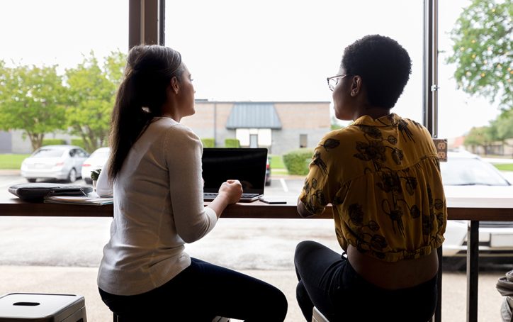 Two women sit at a window counter with laptops, talking and looking outside at parked cars and trees.