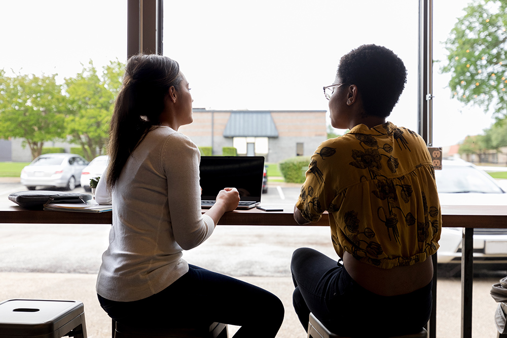 Two women sit at a window counter with laptops, talking and looking outside at parked cars and trees.