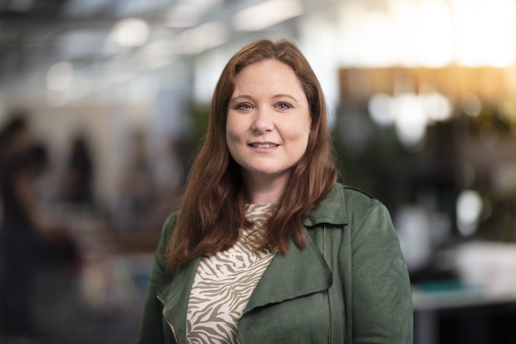 A woman with long brown hair in a green jacket smiles in an office setting with a blurred background.