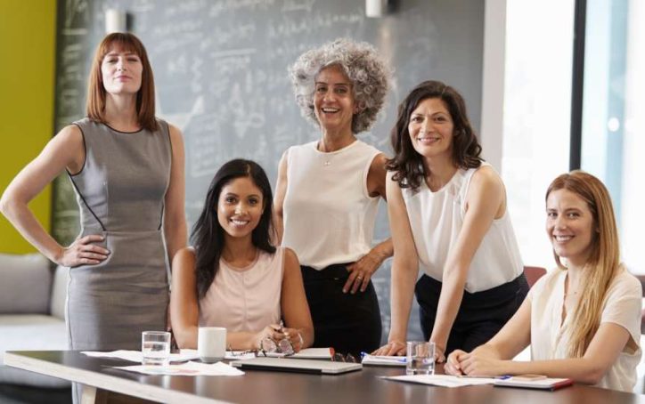 Five women in business attire smiling around a table in a modern office setting with papers and glasses of water.