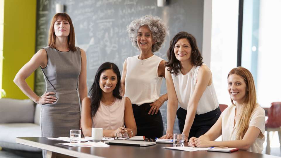 Five women in business attire smiling around a table in a modern office setting with papers and glasses of water.