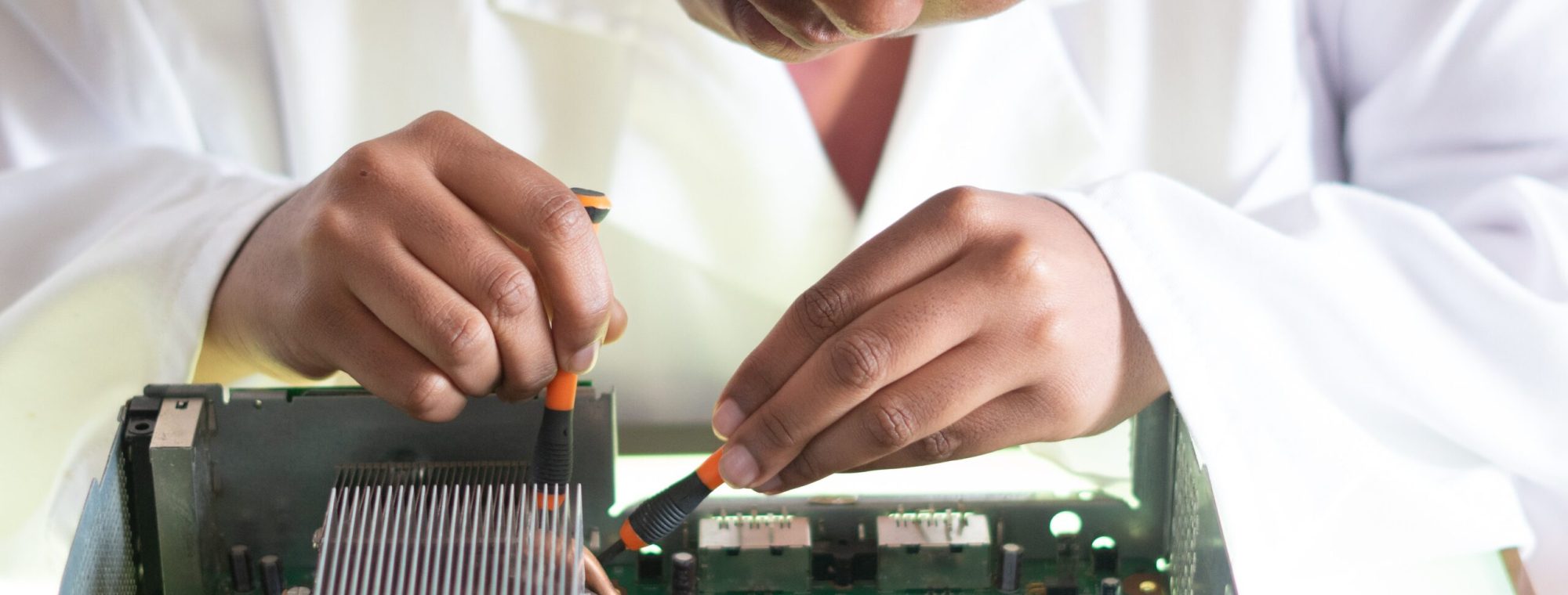 Person in a lab coat repairing a computer motherboard with a screwdriver and a brush.
