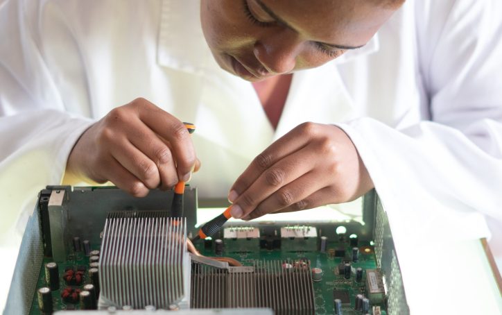 Person in a lab coat repairing a computer motherboard with a screwdriver and a brush.