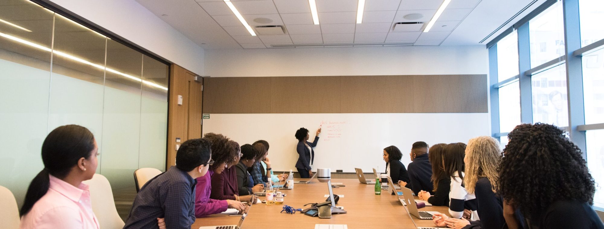 A group of people in a conference room watch a presenter writing on a whiteboard during a meeting.