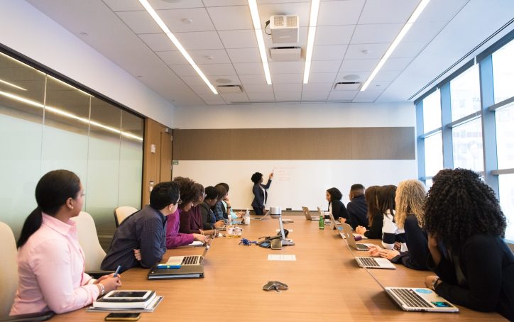 A group of people in a conference room watch a presenter writing on a whiteboard during a meeting.