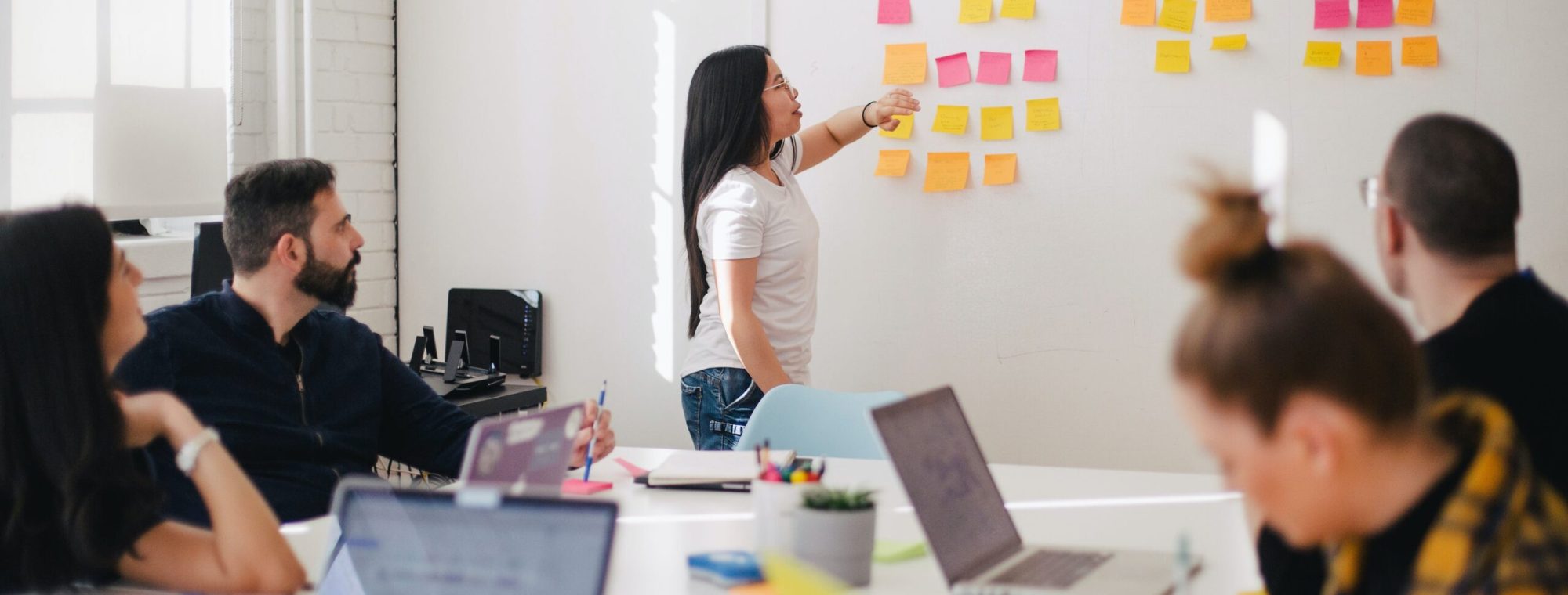 Woman leading a meeting, pointing at sticky notes on a whiteboard, with team members at a table.