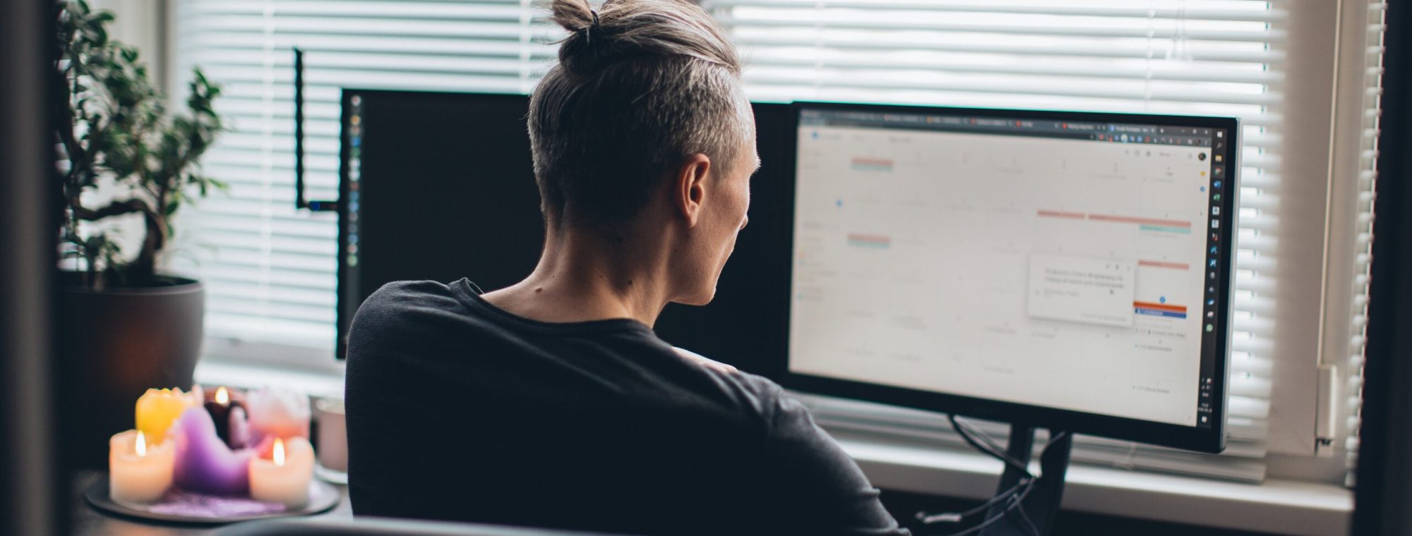 Person with a topknot hairstyle working at a desk with two monitors beside a window with blinds.