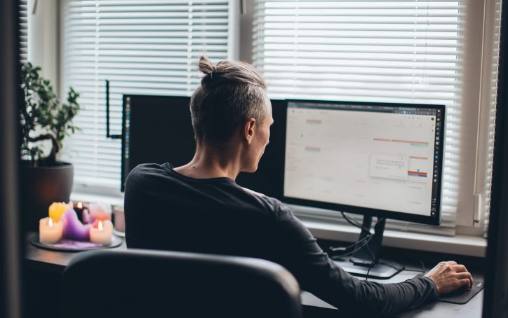 Person with a topknot hairstyle working at a desk with two monitors beside a window with blinds.