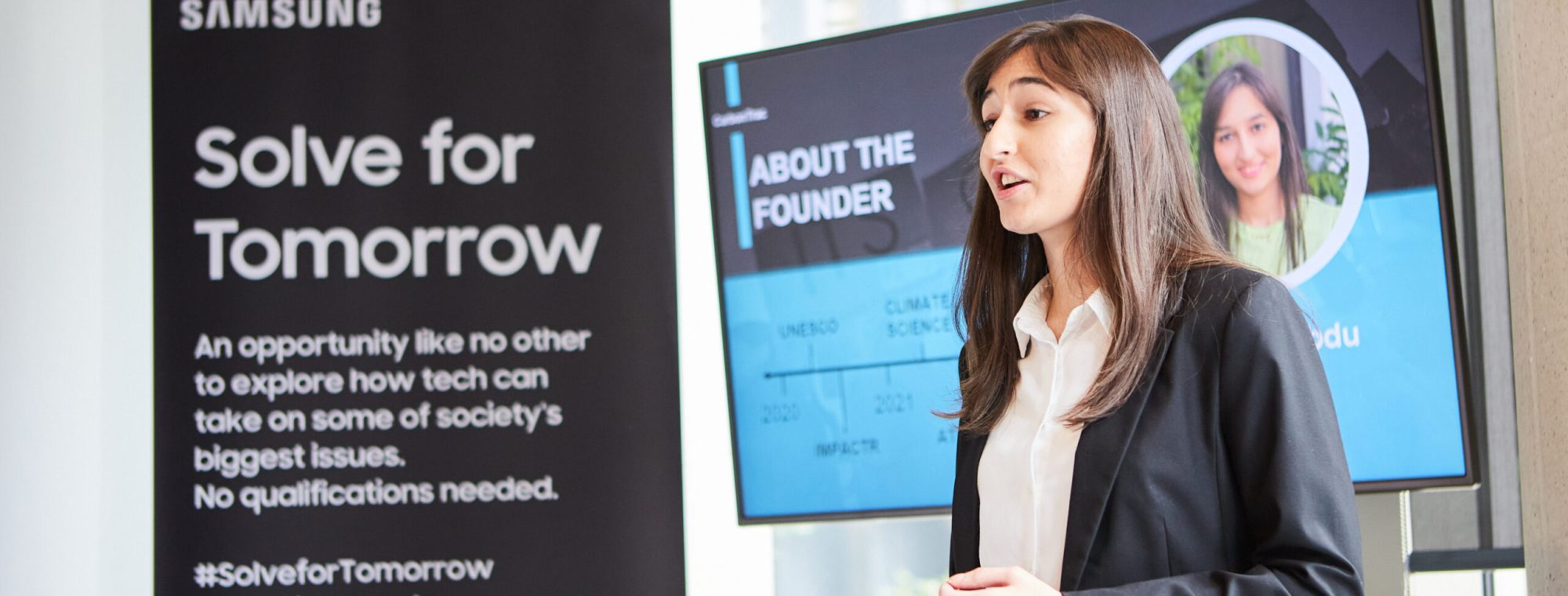 A young woman in business attire presents near a Samsung "Solve for Tomorrow" banner and a screen about the founder.