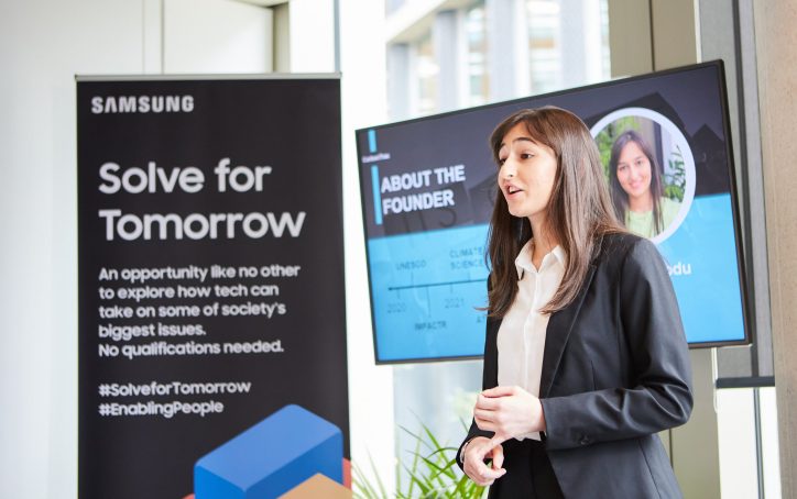 A young woman in business attire presents near a Samsung "Solve for Tomorrow" banner and a screen about the founder.