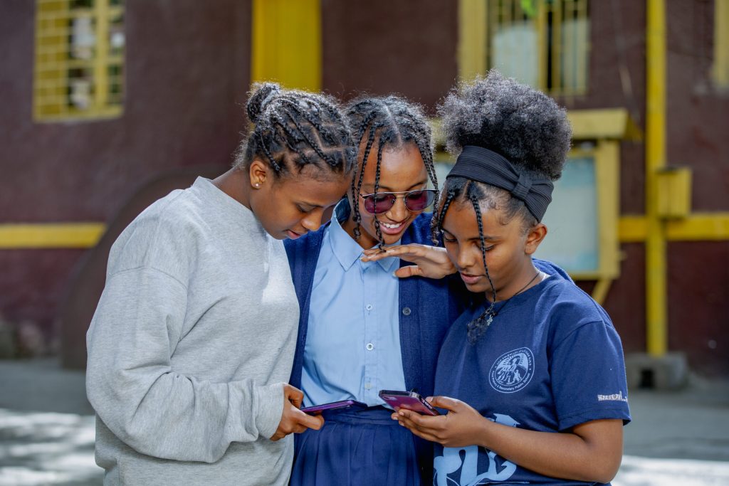 Three girls standing together outdoors, smiling and looking at their phones.