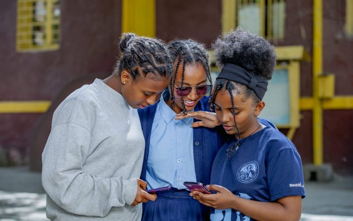 Three girls standing together outdoors, smiling and looking at their phones.