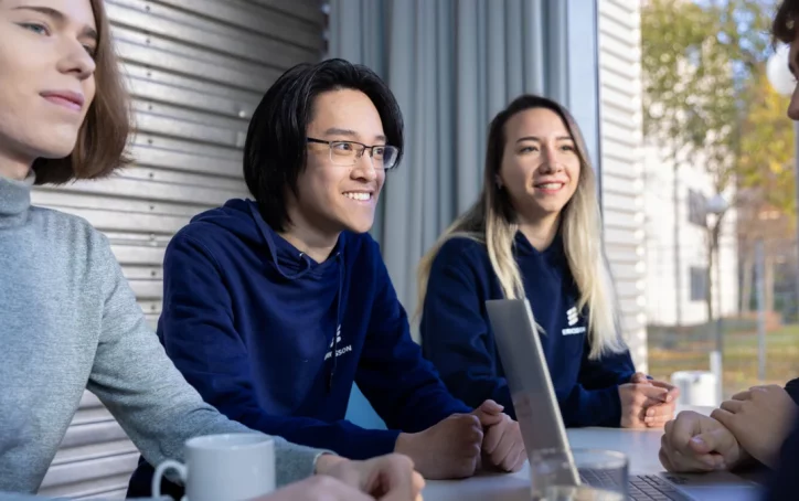 Four people sit together at a table, smiling and talking, with a laptop and drinks in front of them.
