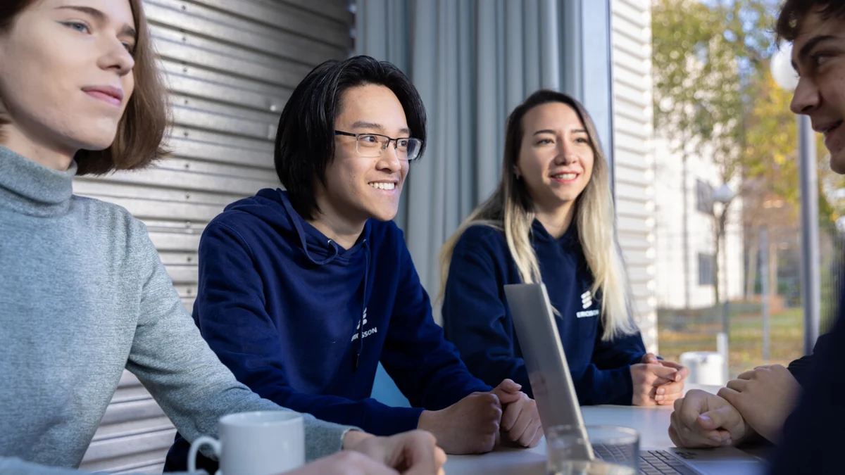 Four people sit together at a table, smiling and talking, with a laptop and drinks in front of them.