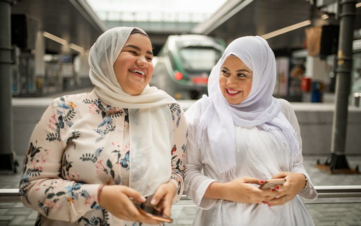 Two women in hijabs smiling and holding phones at a train station platform with a train in the background.