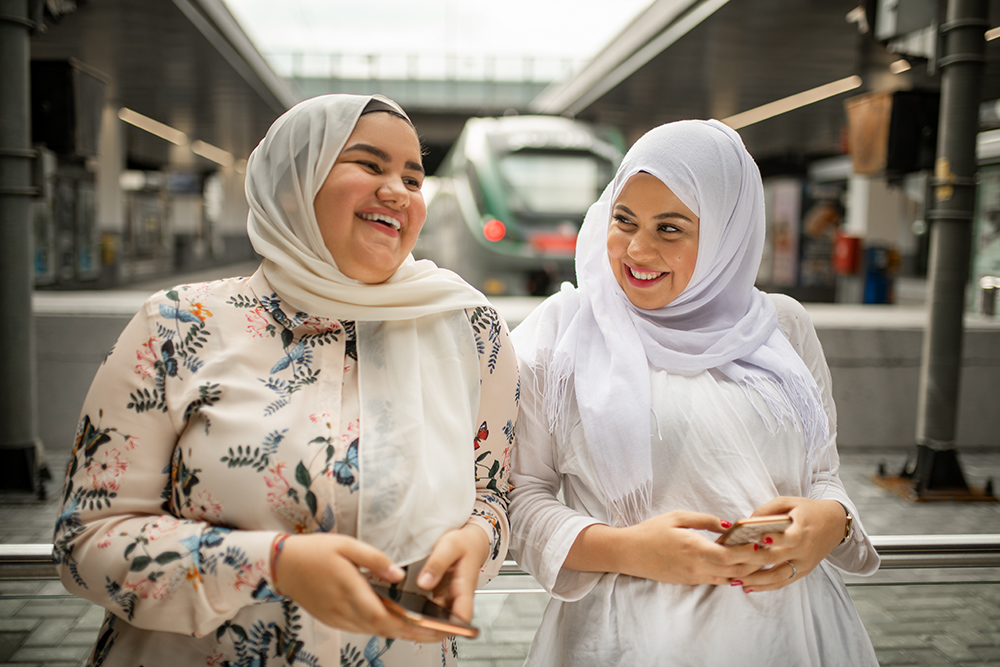 Two women in hijabs smiling and holding phones at a train station platform with a train in the background.