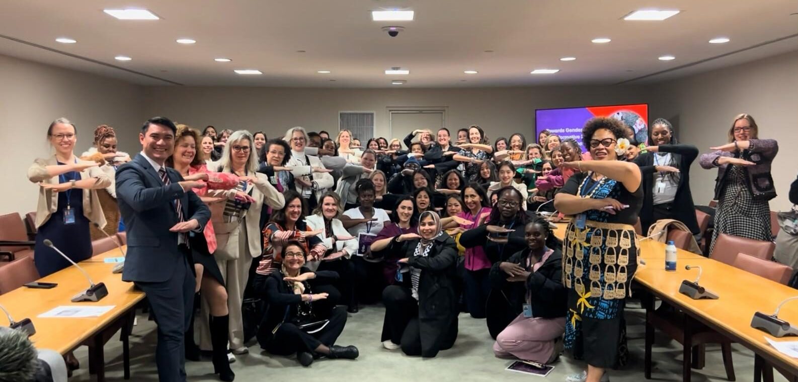 A diverse group of people pose in a conference room, all making an equal sign gesture with their arms.