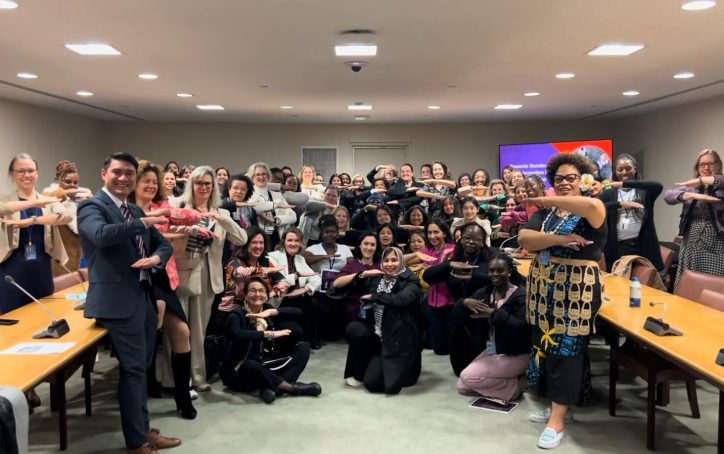 A diverse group of people pose in a conference room, all making an equal sign gesture with their arms.
