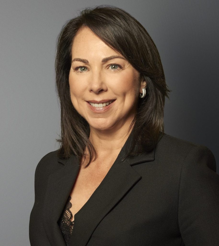 A woman with shoulder-length dark hair in a black blazer smiles at the camera against a gray background.