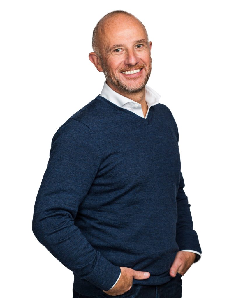 Smiling man with a beard in a blue sweater and white shirt standing with hands in his pockets.