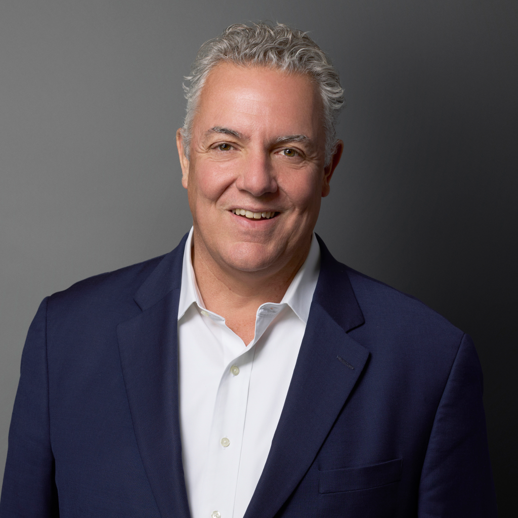 Smiling man with gray hair in a navy blue suit and white shirt against a gray background.