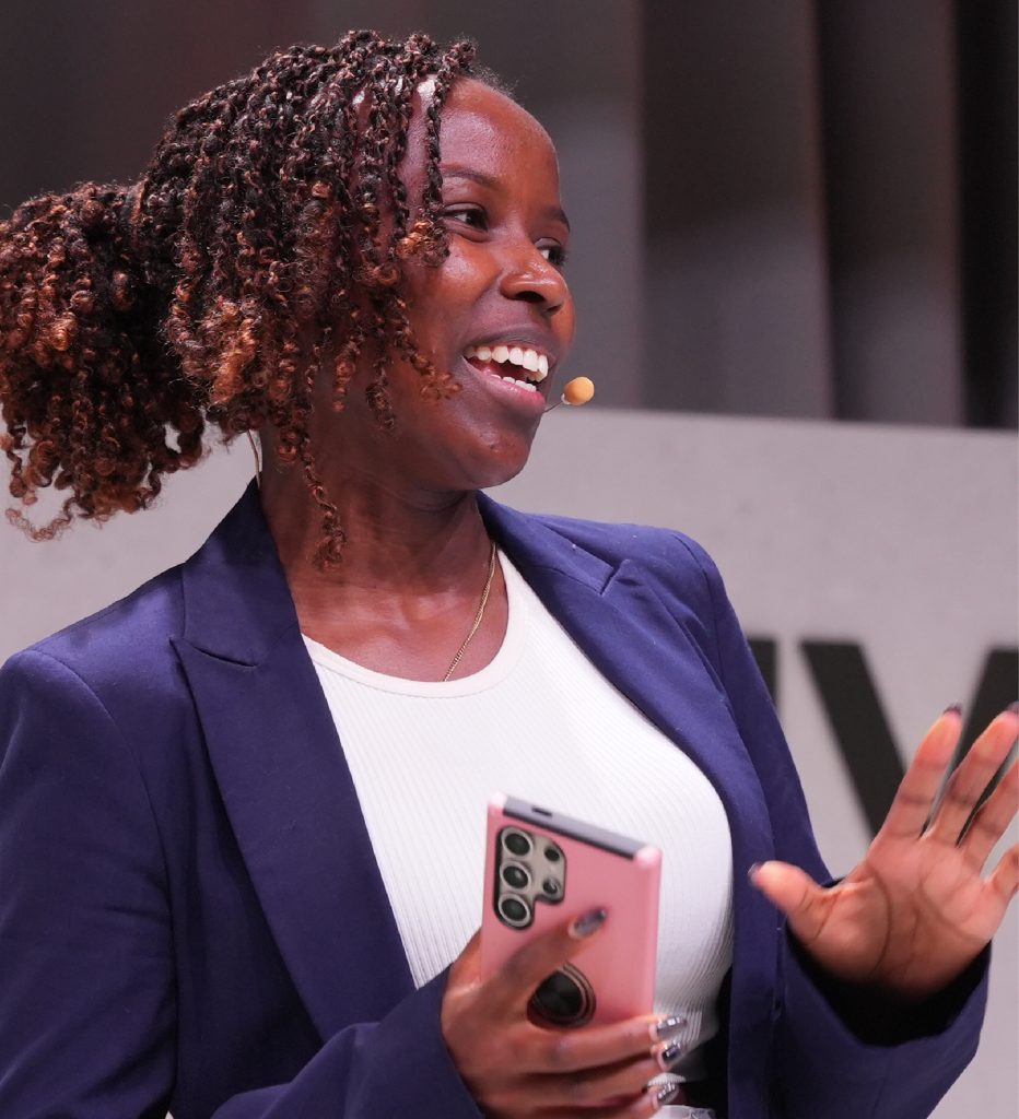 Smiling woman in a blazer speaks on stage with a microphone headset and holds a smartphone in one hand.