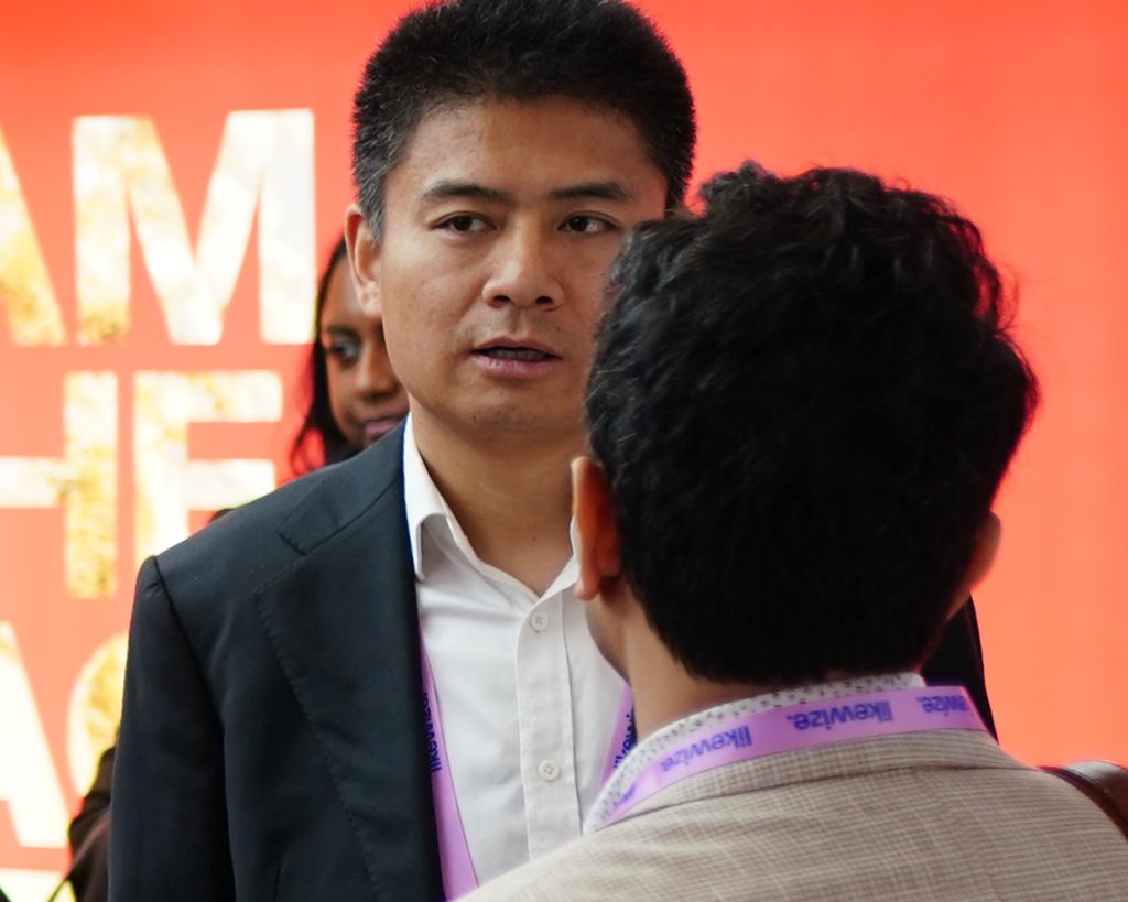 Two men talk at an event; one faces the camera, the other has his back to it, with a red backdrop behind them.