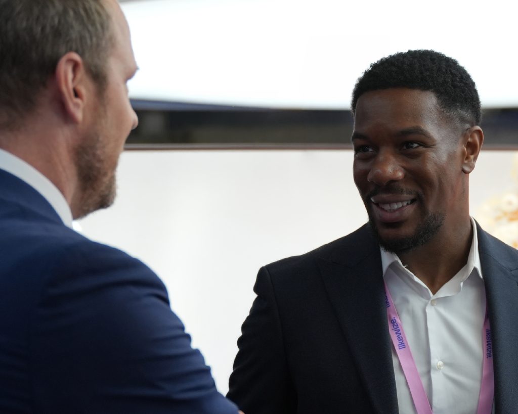 Two men in suits talking and smiling at each other, one wearing a pink lanyard.