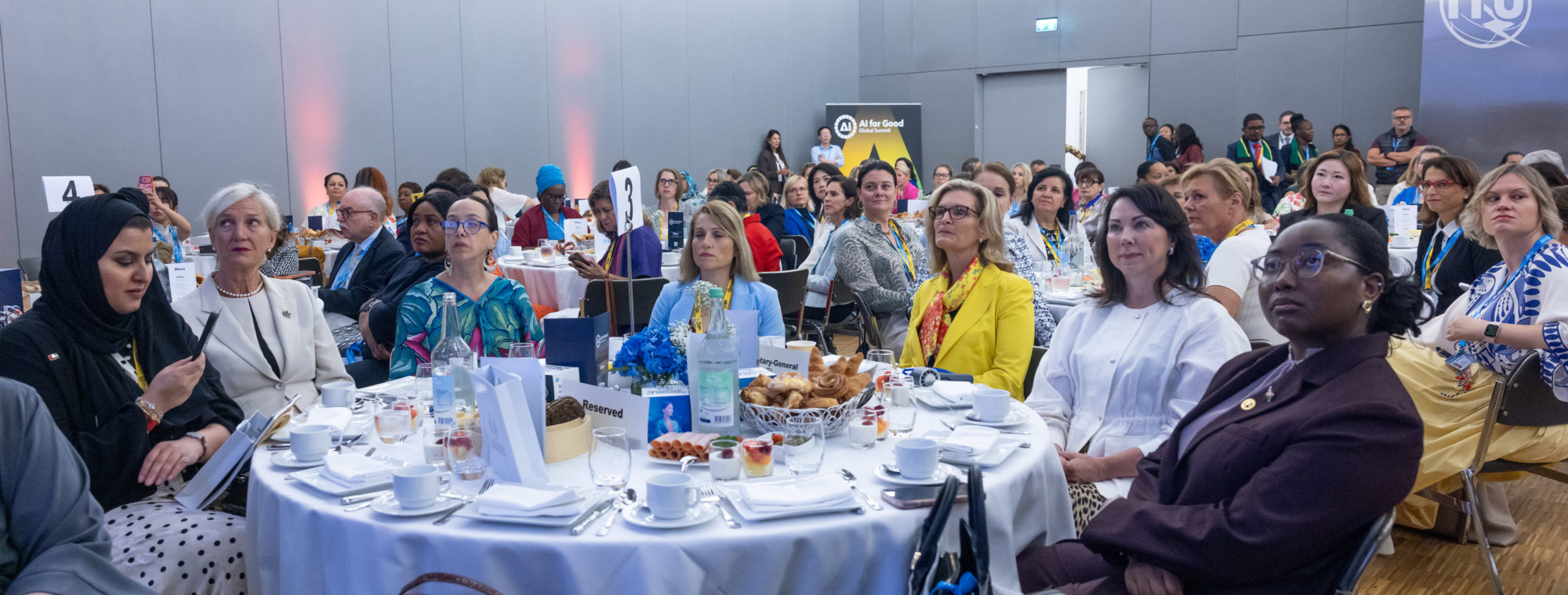 A diverse group of women and a few men sit around round tables, attentively listening at a formal conference. Tables are set with white cloths, tea sets, flowers, and pastries. Attendees wear business or cultural attire in a brightly lit room.