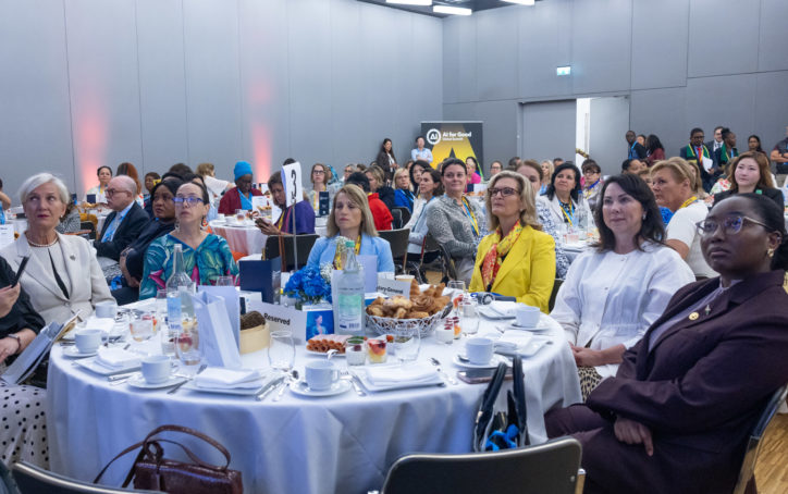 A diverse group of women and a few men sit around round tables, attentively listening at a formal conference. Tables are set with white cloths, tea sets, flowers, and pastries. Attendees wear business or cultural attire in a brightly lit room.