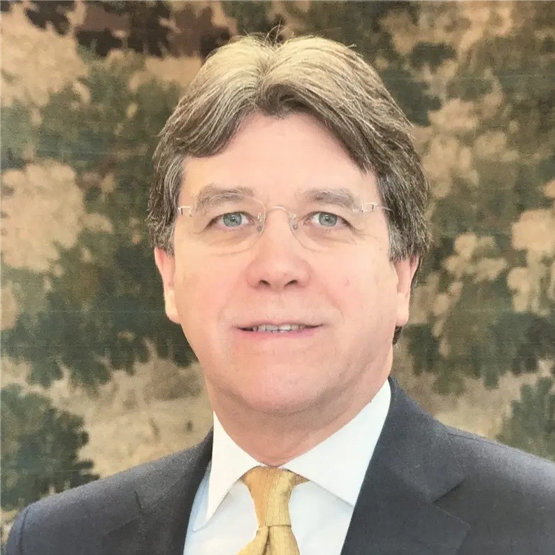 A middle-aged man with glasses, grayish-brown hair, and a slight smile wears a dark suit, light shirt, and yellow tie. The background is a blurred green and beige tapestry.