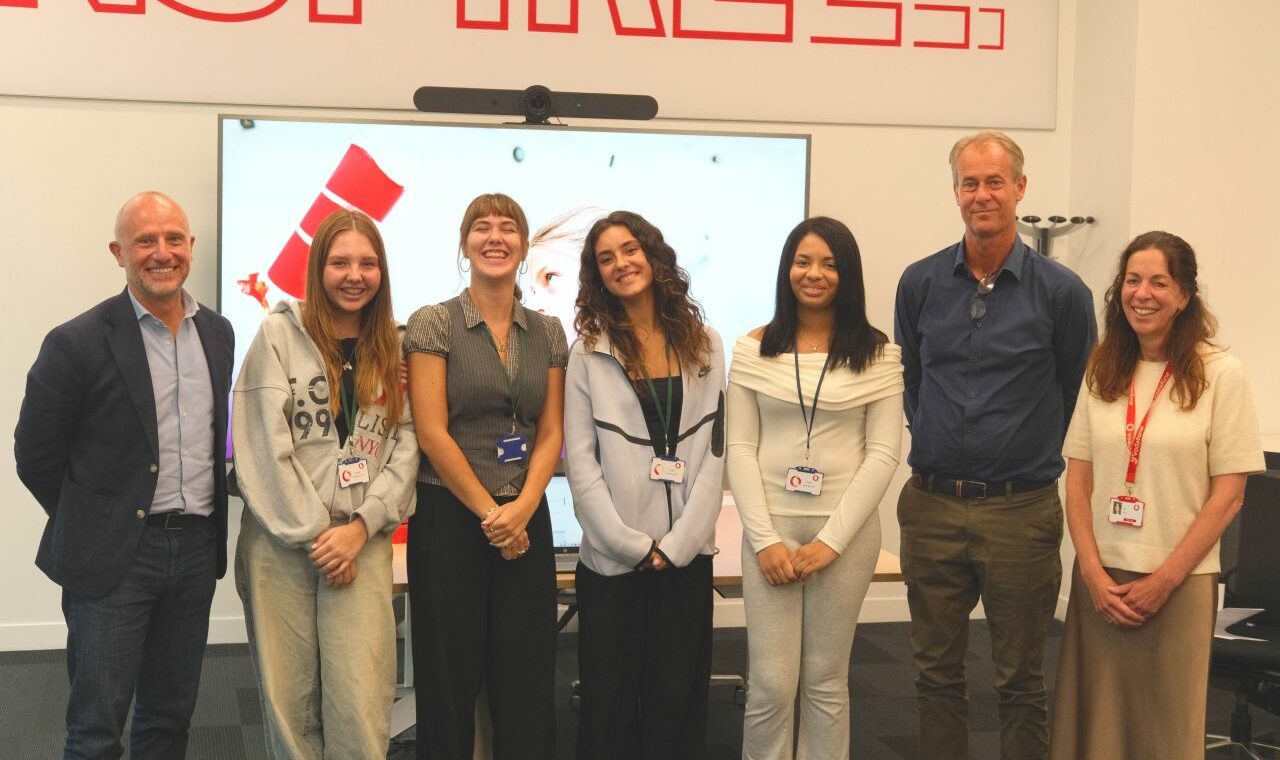 Seven people stand indoors in a line, smiling at the camera. Four women and three men wear smart-casual clothes. Behind them is a TV screen, part of a red word “INSPIRE” on the wall, and a whiteboard. All wear lanyards or badges.