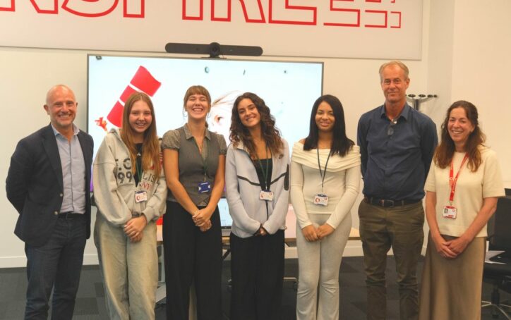 Seven people stand indoors in a line, smiling at the camera. Four women and three men wear smart-casual clothes. Behind them is a TV screen, part of a red word “INSPIRE” on the wall, and a whiteboard. All wear lanyards or badges.