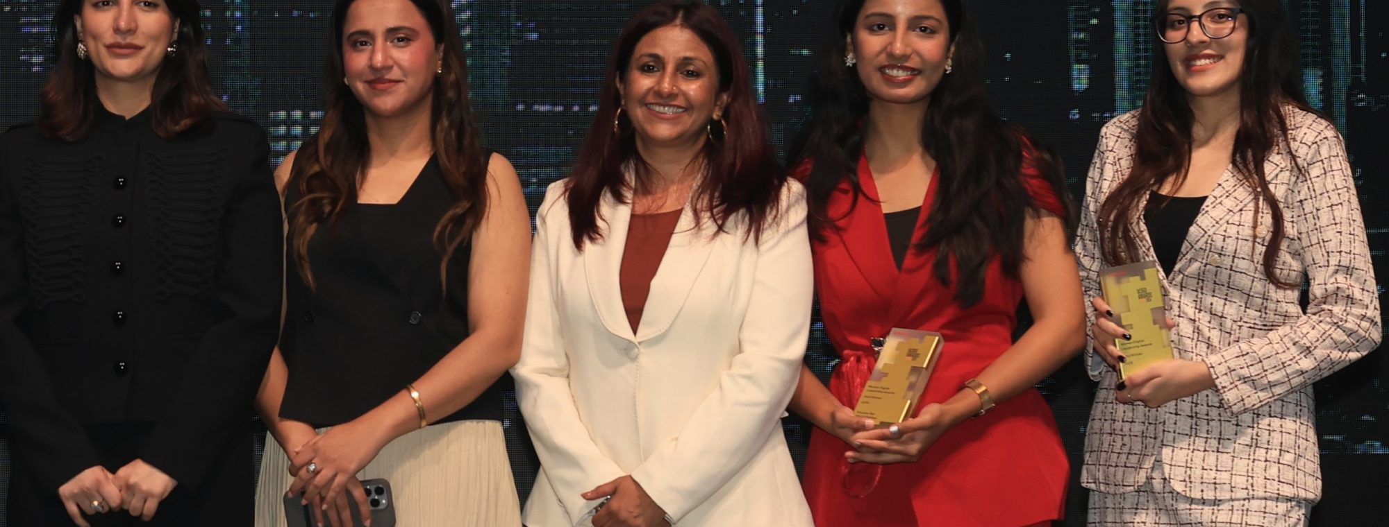 Five women stand side by side, smiling at the camera at the Women Digital Leadership Awards 2026—an event celebrating women shaping the future of AI. Two hold awards. Outfits range from a black suit, a white blazer and pants, a black sleeveless top with a cream pleated skirt, a red pantsuit, and a white plaid dress with black boots.