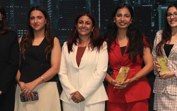 Five women stand side by side, smiling at the camera at the Women Digital Leadership Awards 2026—an event celebrating women shaping the future of AI. Two hold awards. Outfits range from a black suit, a white blazer and pants, a black sleeveless top with a cream pleated skirt, a red pantsuit, and a white plaid dress with black boots.