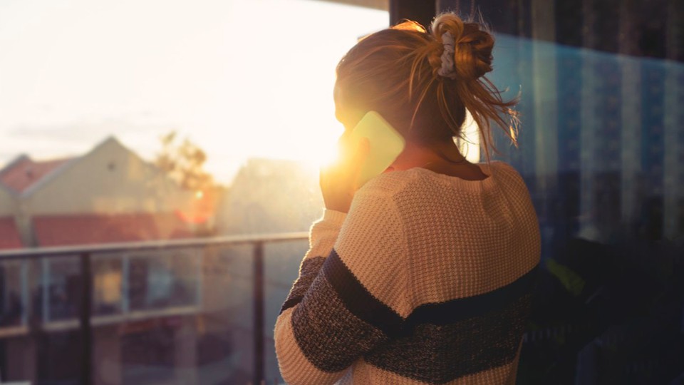 A person stands facing a window, talking on a light-colored smartphone. The bright, warm glow of the setting sun frames them from behind.